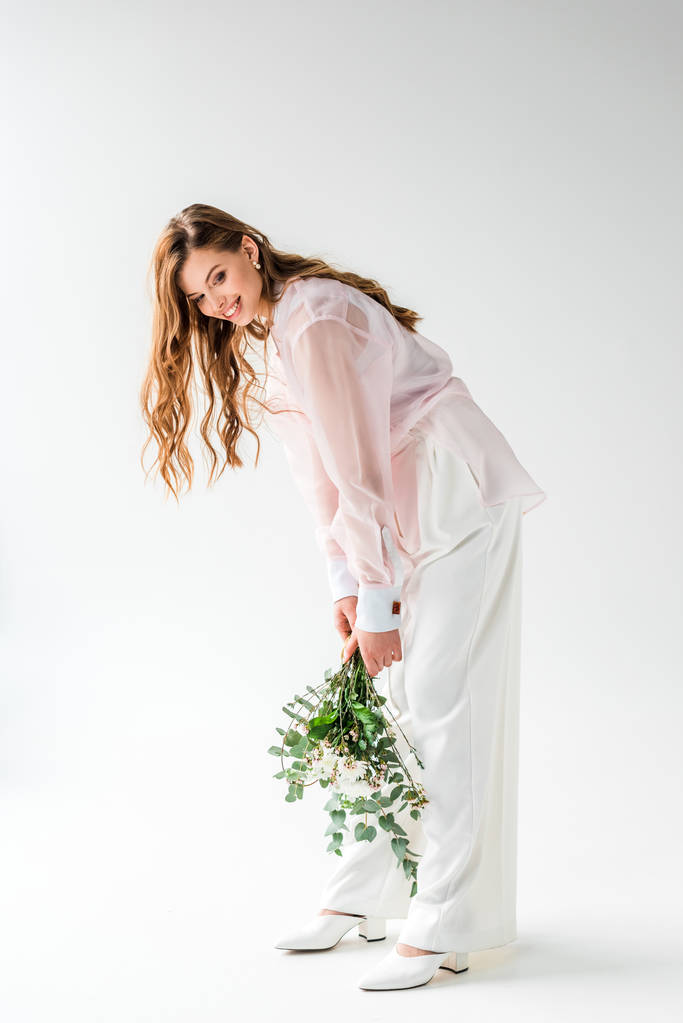 happy and stylish young woman holding flowers with eucalyptus leaves on white  - Photo, Image
