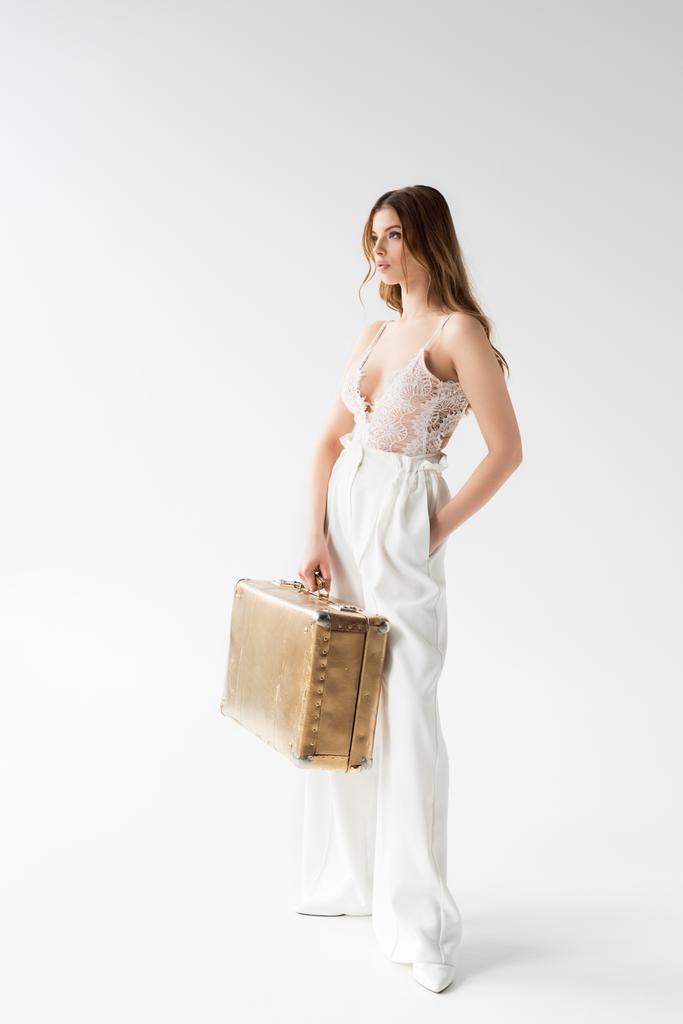 стильная симпатичная девушка, держащая чемодан, стоя с рукой в кармане на белом
  - Фото, изображение