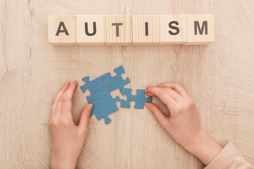 частичный вид женских рук с голубыми головоломками на деревянном столе рядом с деревянными кубиками с надписью аутизма
 - Фото, изображение