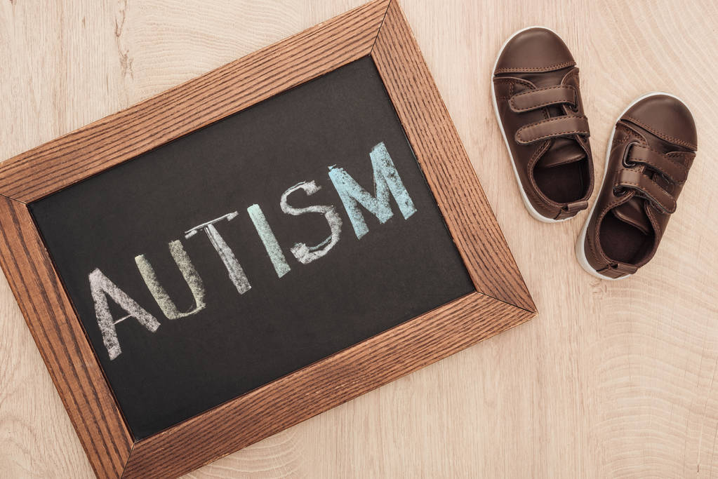 bovenaanzicht van autisme letters geschreven op een schoolbord in de buurt van kinderen bruin sneakers op houten oppervlak - Foto, afbeelding