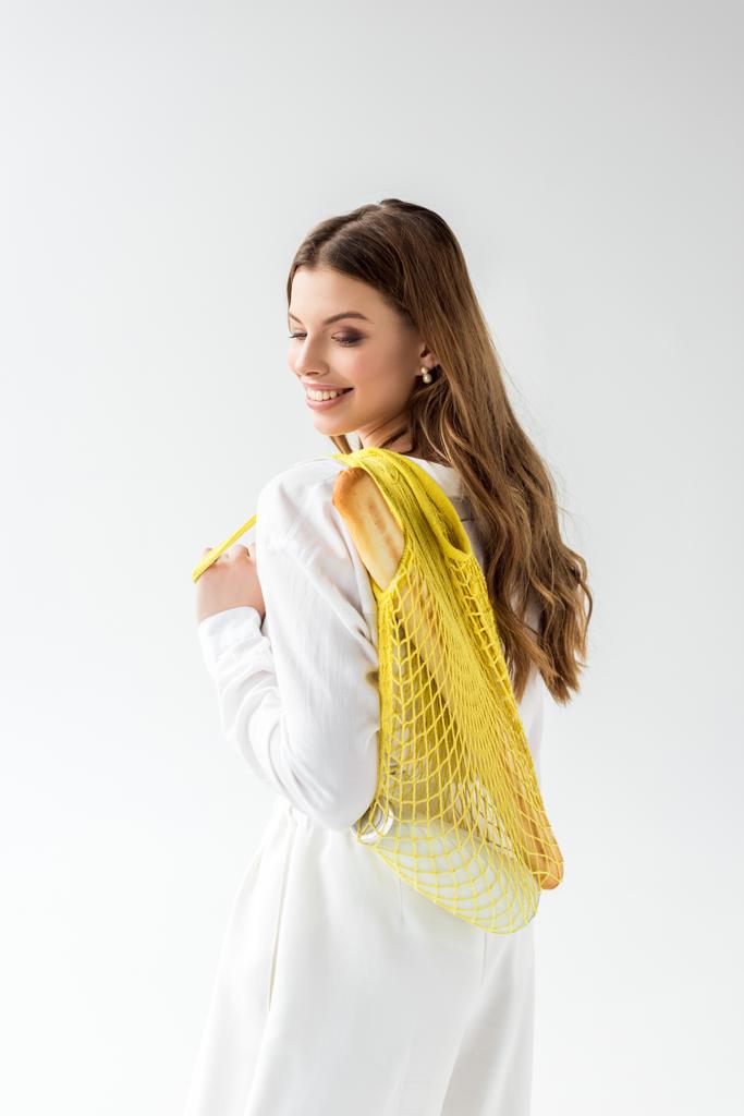 cheerful young woman holding yellow string bag with baguette and bottle of milk on white  - Photo, Image