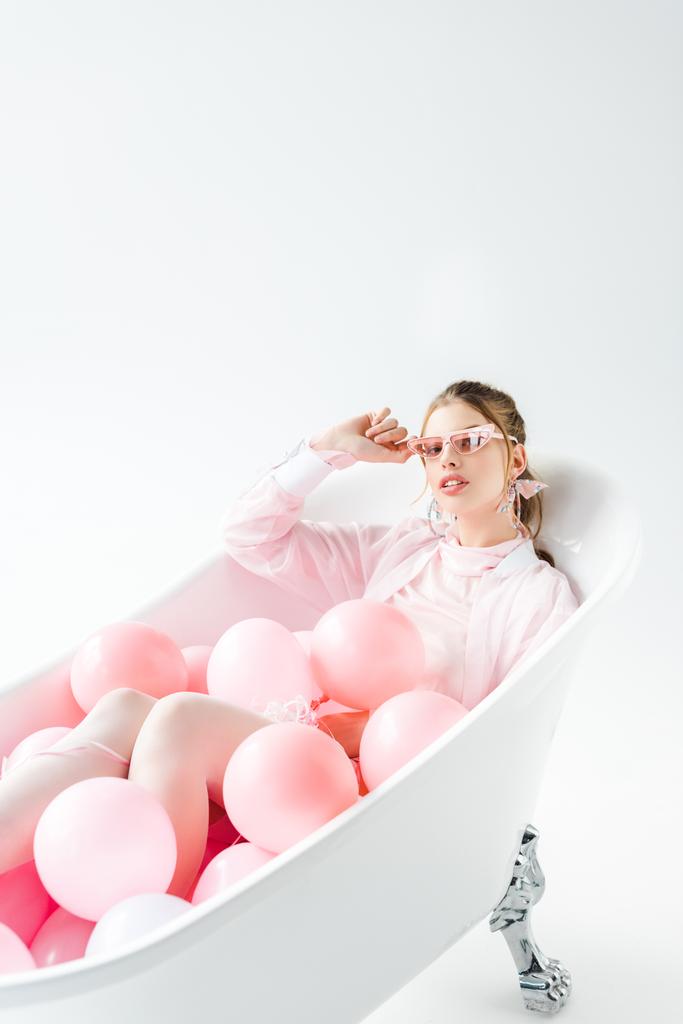 elegante giovane donna sdraiata nella vasca da bagno con palloncini d'aria rosa e occhiali da sole toccanti su bianco
  - Foto, immagini