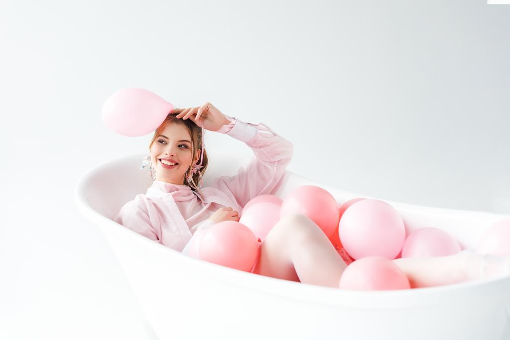 happy young woman lying in bathtub and holding pink air balloon on white  - Photo, Image