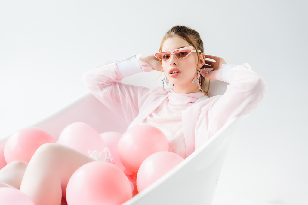 mujer joven con estilo en gafas de sol tumbado en la bañera con globos de aire de color rosa en blanco
  - Foto, imagen
