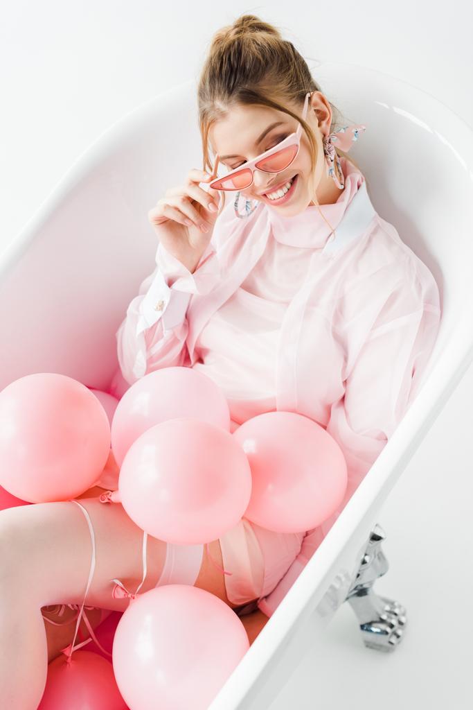 bovenaanzicht van vrolijk meisje aanraken zonnebril liggend in badkuip met roze lucht ballonnen op wit  - Foto, afbeelding