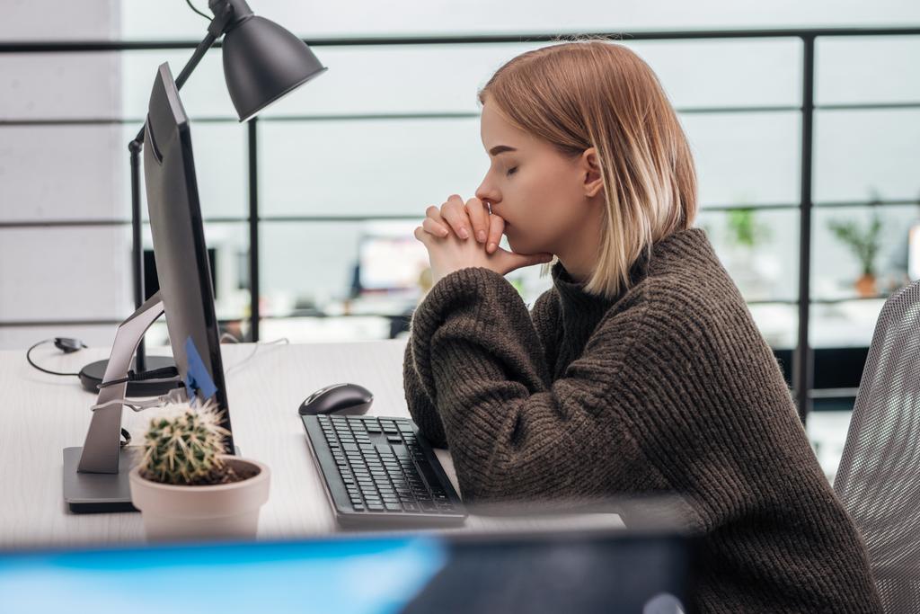 избирательный фокус расстроенной девушки, сидящей на рабочем месте с сжатыми руками и закрытыми глазами в современном офисе
 - Фото, изображение