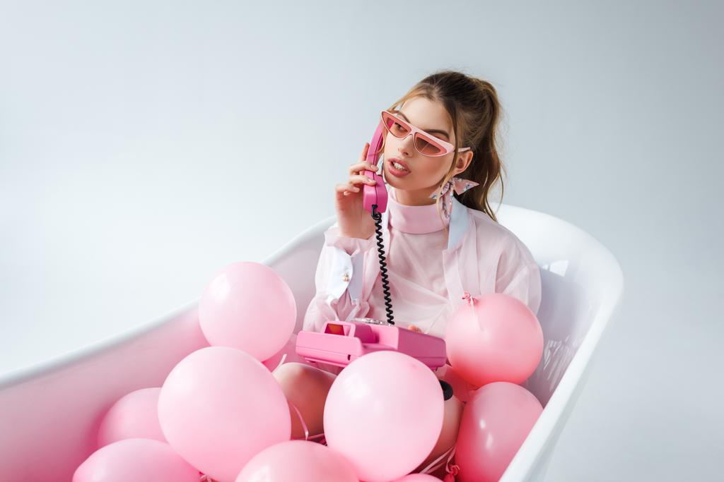junge Frau mit Sonnenbrille, die in der Badewanne liegt und mit rosa Luftballons auf weißem Grund telefoniert  - Foto, Bild
