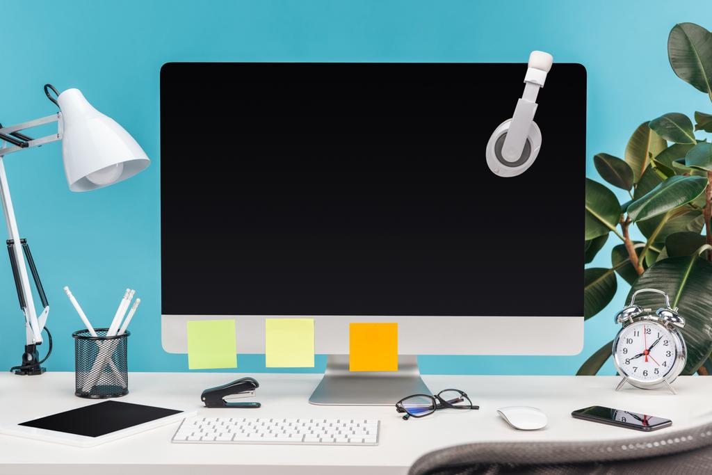 lieu de travail avec écouteurs et notes collantes vides sur écran d'ordinateur, lampe et papeterie sur table blanche sur fond bleu
 - Photo, image