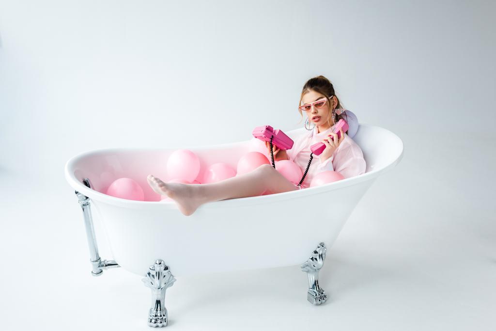 девушка в солнцезащитных очках смотрит на розовый ретро-телефон, пока лежит в ванне с воздушными шарами на белом
  - Фото, изображение