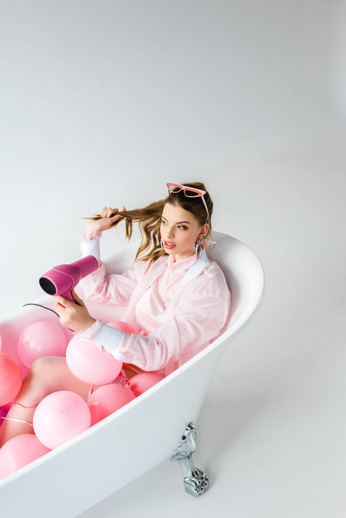 приваблива дівчина використовує фен, лежачи у ванні з рожевими повітряними кульками на сірому
  - Фото, зображення