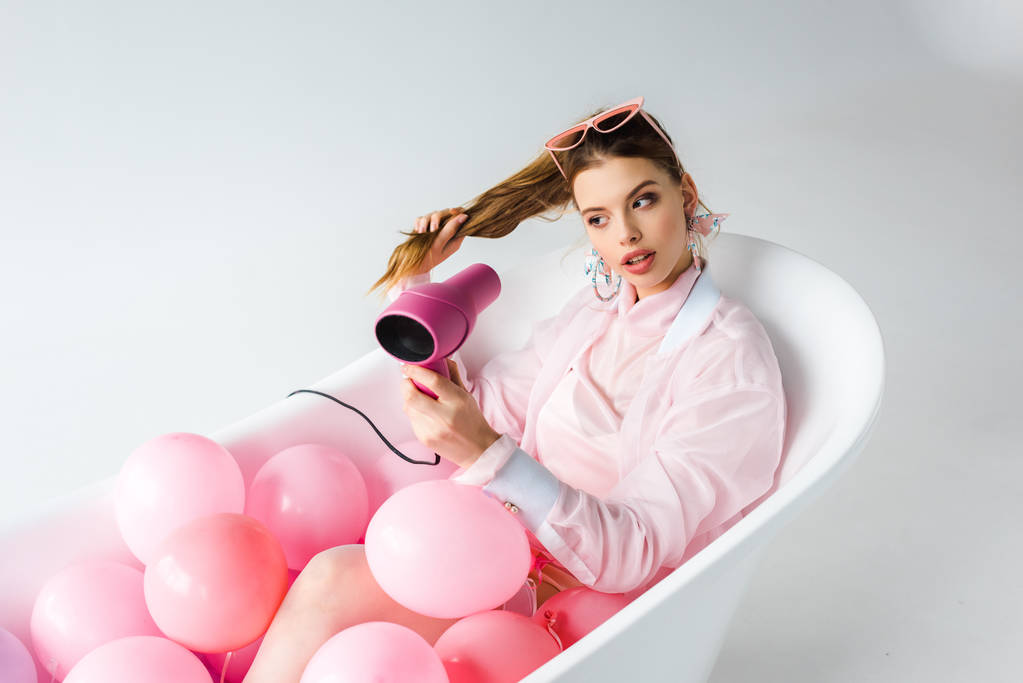 приваблива дівчина використовує фен, лежачи у ванні з рожевими повітряними кульками на білому
  - Фото, зображення