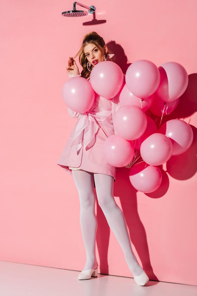 όμορφη νεαρή γυναίκα κρατώντας αέρα μπαλόνια ενώ αγγίζοντας τα μαλλιά σε ροζ - Φωτογραφία, εικόνα