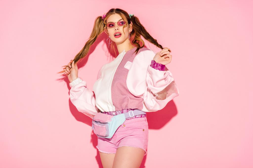 verbaasd stijlvolle meisje in zonnebrillen en taille tas aanraken van haren op roze - Foto, afbeelding