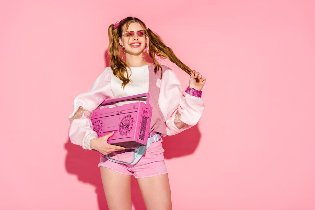 happy stylish girl in sunglasses holding boombox and touching hair on pink - Photo, Image