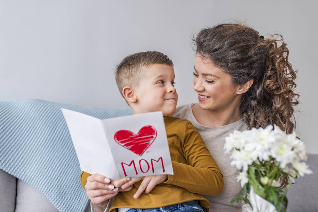 Lindo chico con tarjeta de felicitación sentado con la madre sosteniendo flores
 - Foto, Imagen