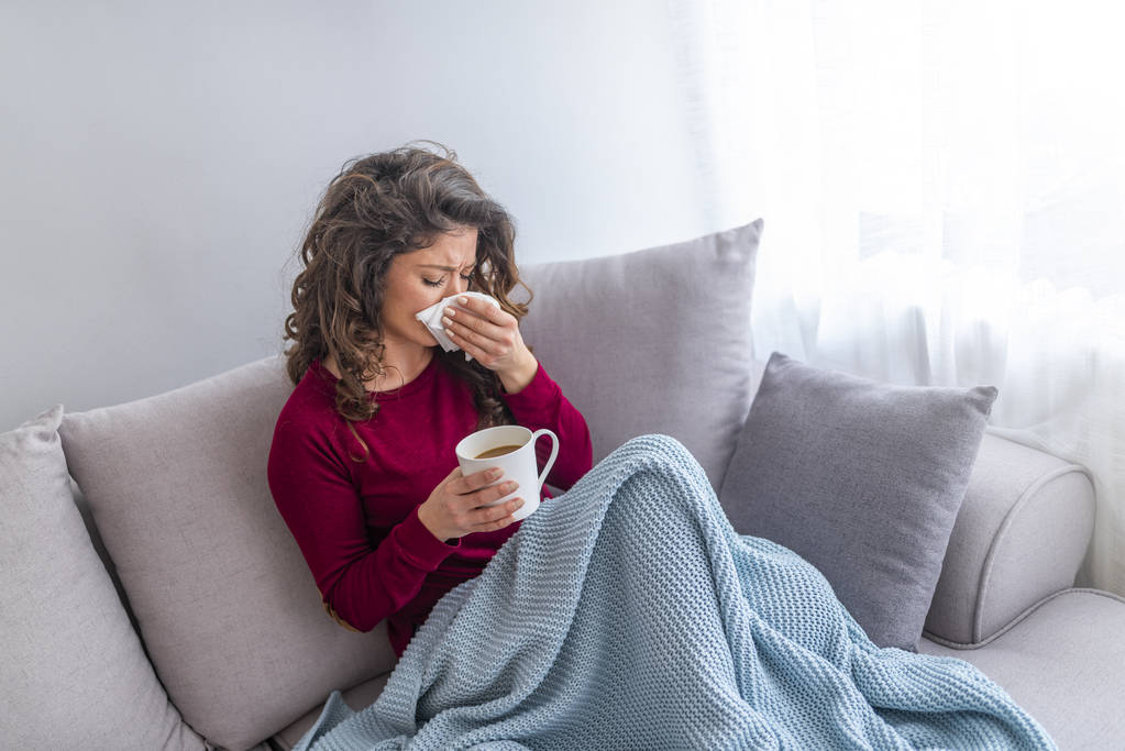 Young girl sick in bed with temperature sneezing into tissue and holding fever drinking cup of warm tea - Photo, Image