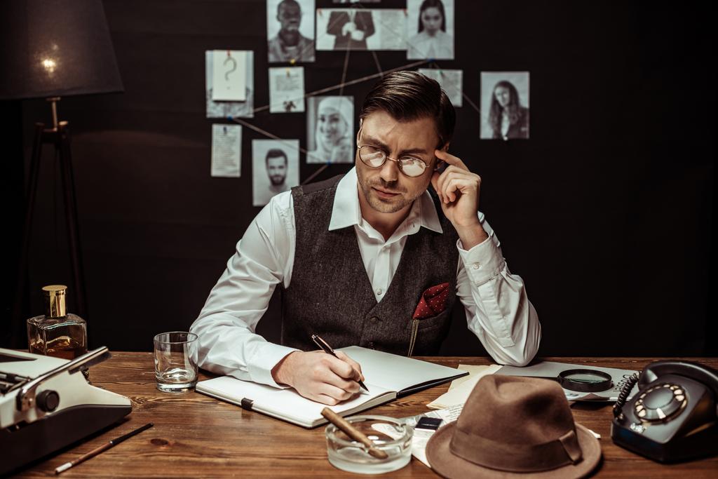 Pensive detective in glasses sitting at table and writing in notebook - Photo, Image