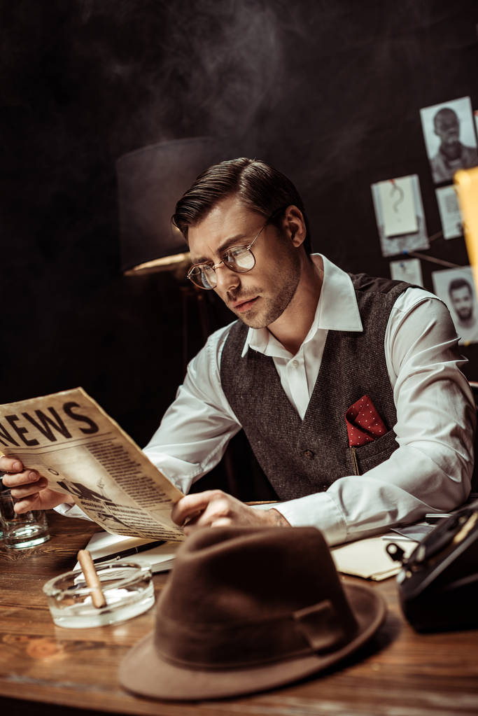Concentrated detective in glasses reading newspaper in dark office - Photo, Image