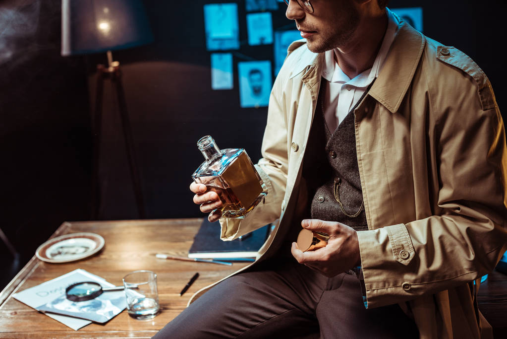 Περικομμένη θέα του ντετέκτιβ σε καμπαρντίνα κάθεται στο τραπέζι και κρατώντας ένα μπουκάλι κονιάκ - Φωτογραφία, εικόνα
