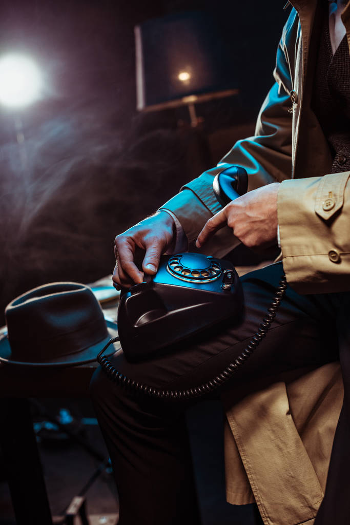 Μερική θέα του ανθρώπου σε καμπαρντίνα χρησιμοποιώντας το τηλέφωνο σε σκοτεινό γραφείο - Φωτογραφία, εικόνα