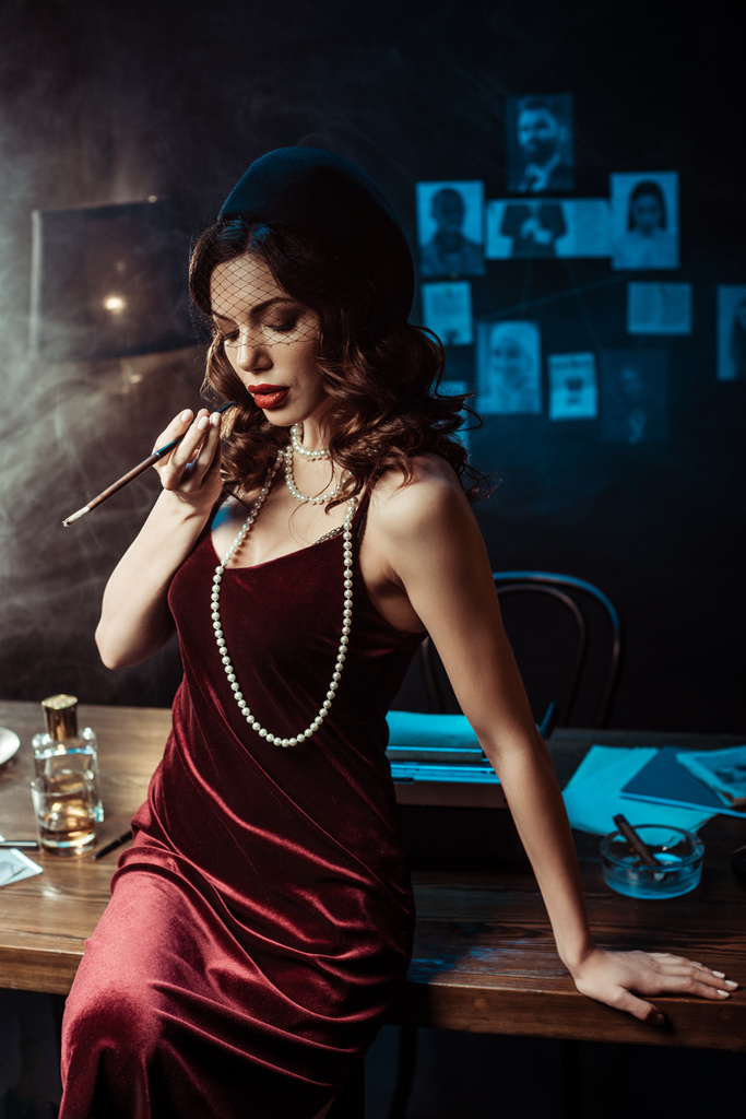 Женщина в платье сидит на столе и держит мундштук в темном кабинете
 - Фото, изображение