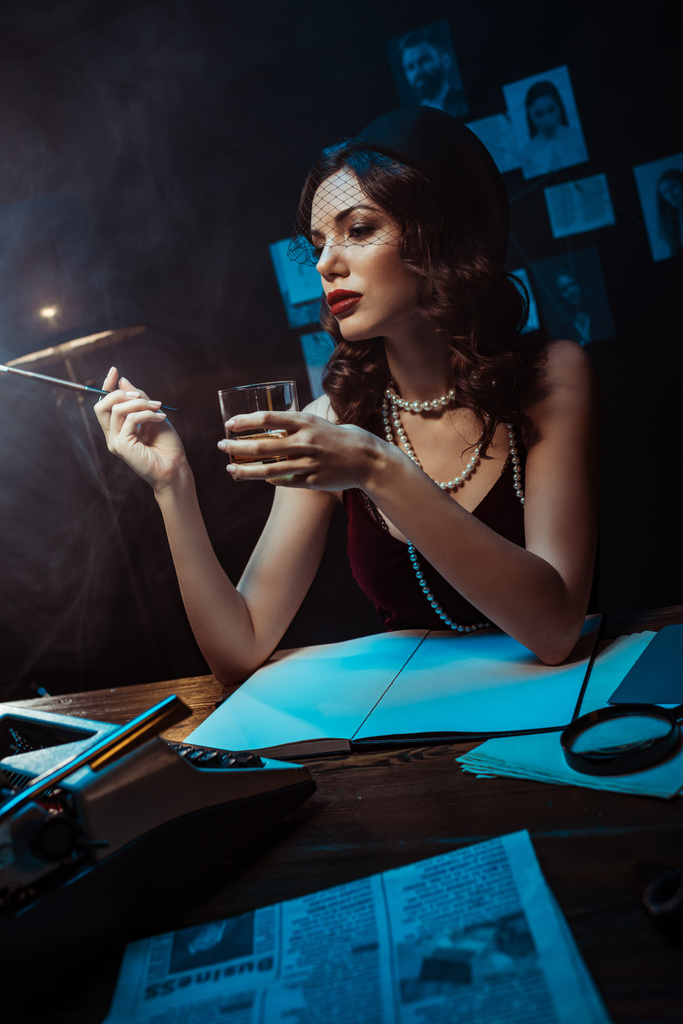 Karanlık ofiste aksesuarı şapka tutan ağız parçası ve konyak cam çekici kadın - Fotoğraf, Görsel