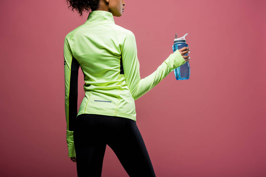 Achteraanzicht van Afro-Amerikaanse Sportvrouw in track jas met sport fles geïsoleerd op brown - Foto, afbeelding