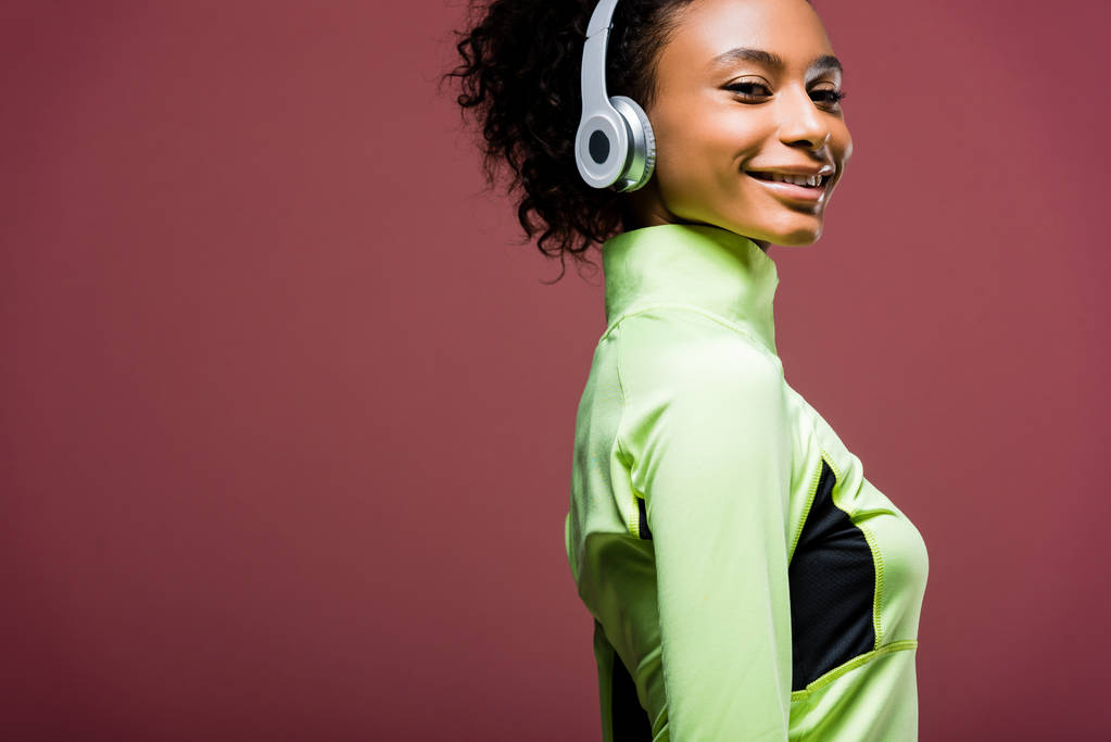 beautiful smiling african american sportswoman in headphones looking at camera isolated on brown with copy space - Photo, Image