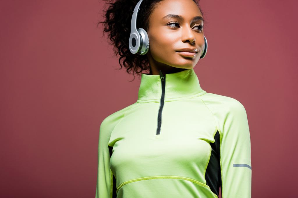 bela esportista afro-americana em fones de ouvido olhando para longe isolado em marrom com espaço de cópia
 - Foto, Imagem
