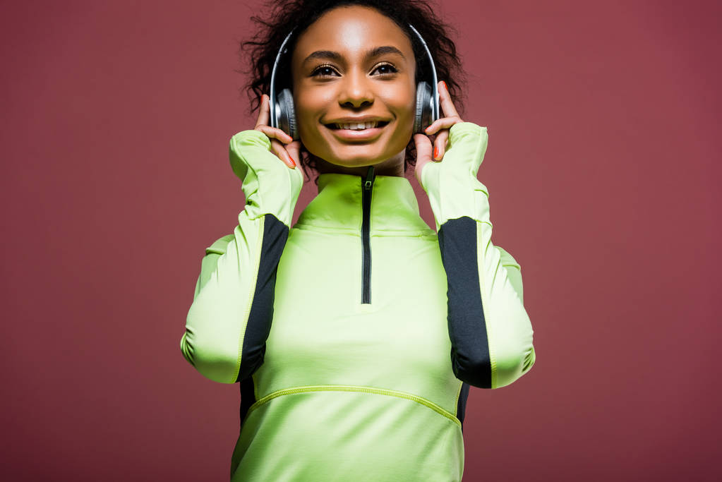 bela sorridente Africano americano desportista em fones de ouvido e jaqueta de pista isolado em marrom
 - Foto, Imagem