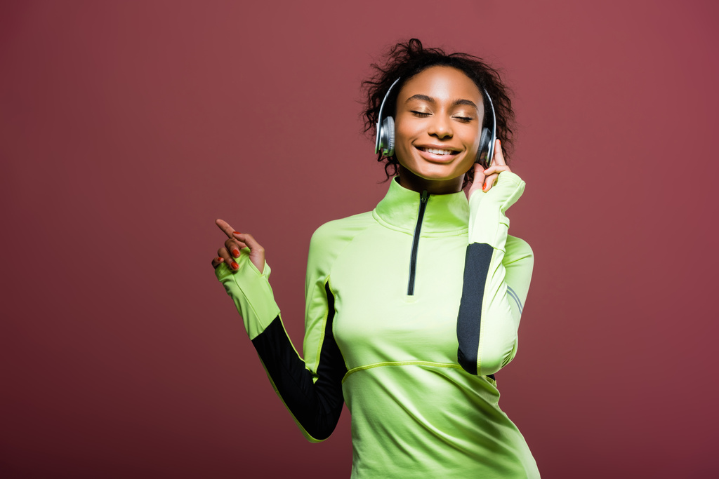 krásné usmívající se africká americká sportovkyně v sluchátka poslech hudby s očima uzavřené izolované na hnědé - Fotografie, Obrázek