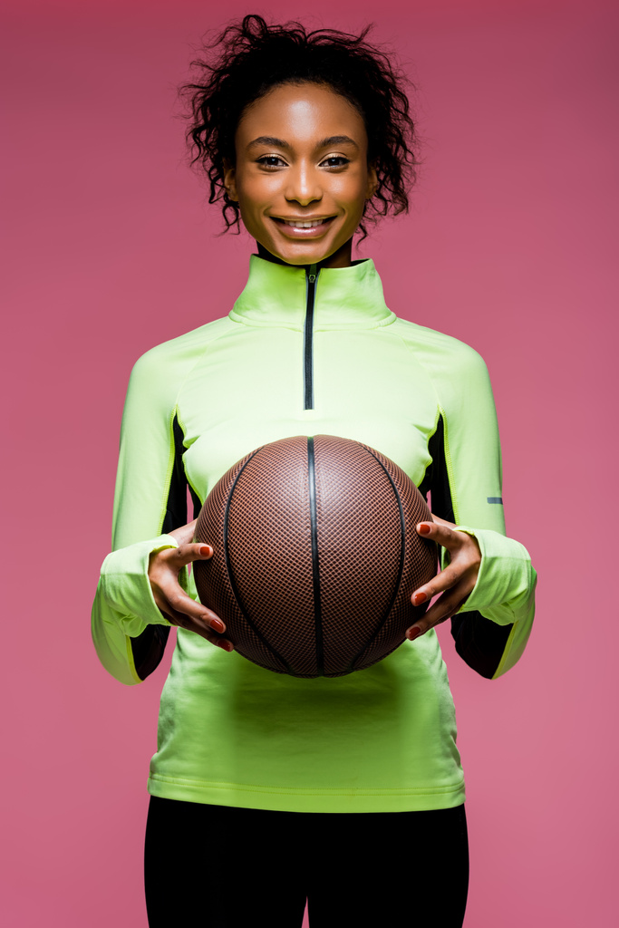 カメラ目線、ピンクに分離されたバスケット ボールを保持している美しいの笑顔のアフリカ系アメリカ人のスポーツウーマン - 写真・画像