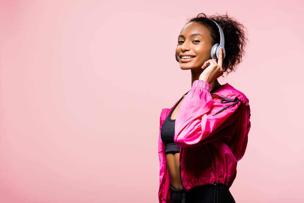 schöne lächelnde afrikanisch-amerikanische Sportlerin mit Kopfhörer, die in die Kamera schaut, isoliert auf rosa mit Kopierraum - Foto, Bild