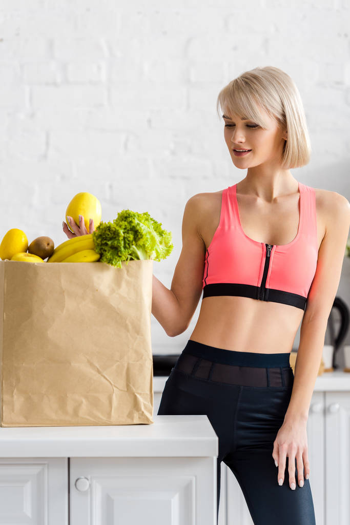 cheerful blonde woman in sportswear looking at paper bag with groceries - Photo, Image