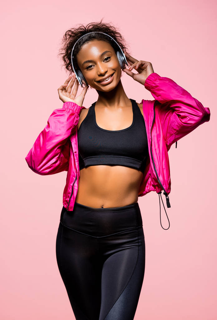 красива афроамериканська спортсменка в навушниках слухає музику і дивиться на камеру ізольовано на рожевому
 - Фото, зображення