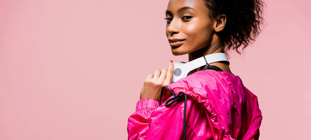 fotografía panorámica de la hermosa deportista afroamericana con auriculares mirando a la cámara aislada en rosa
 - Foto, Imagen