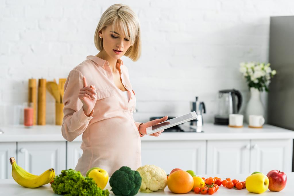 attraente donna incinta con tablet digitale e guardando frutta e verdura in cucina
  - Foto, immagini