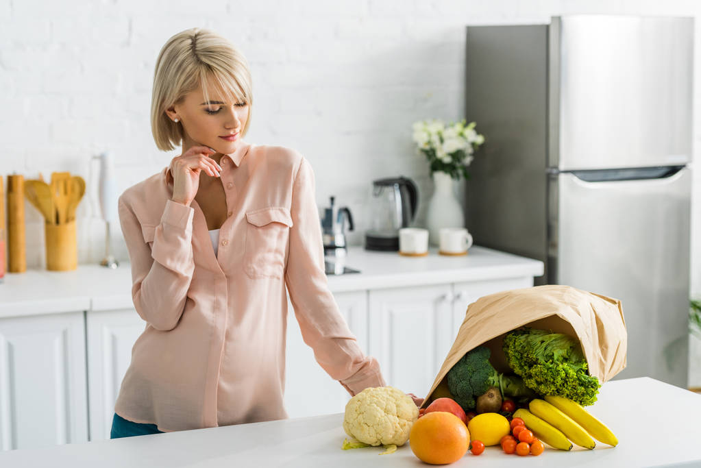 ξανθιά έγκυος γυναίκα κοιτάζοντας νόστιμα λαχανικά και τα φρούτα κοντά σε χάρτινη σακούλα - Φωτογραφία, εικόνα