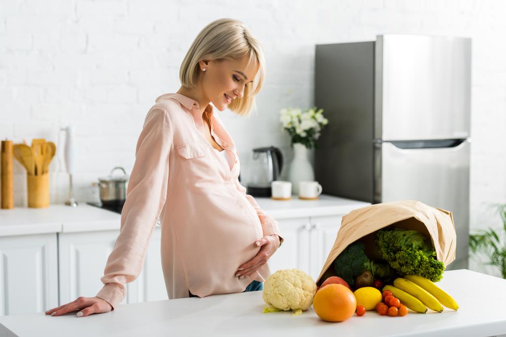 atractiva mujer rubia y embarazada mirando comestibles cerca de bolsa de papel
 - Foto, imagen