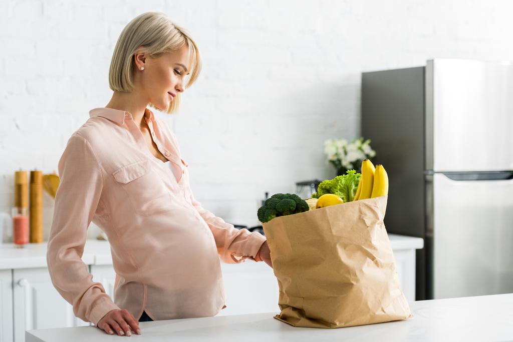 aantrekkelijke blonde en zwangere vrouw te kijken naar papieren zak in de keuken - Foto, afbeelding
