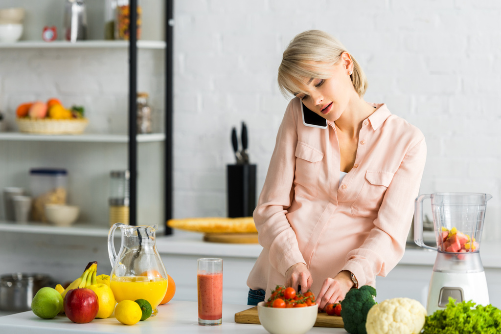 блондинка вагітна жінка говорить на смартфоні на кухні біля фруктів і овочів
  - Фото, зображення