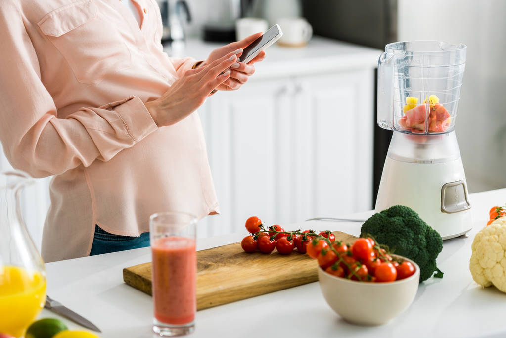vista recortada de la mujer embarazada usando teléfono inteligente en la cocina cerca de tomates cherry en la tabla de cortar
 - Foto, Imagen