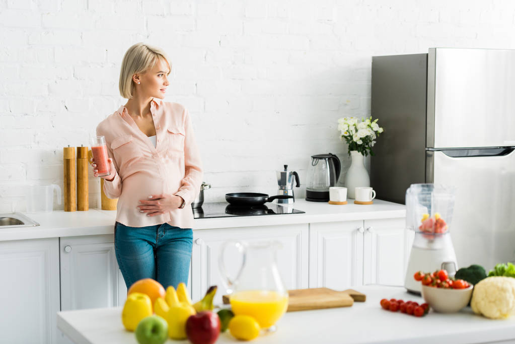 blonde zwangere vrouw bedrijf glas smoothie in de keuken  - Foto, afbeelding