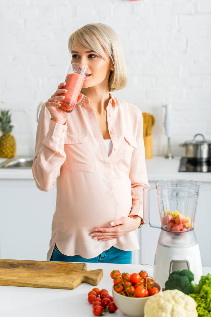 魅力的なブロンドの妊婦は、キッチンで野菜の近くにスムージーを飲みます  - 写真・画像