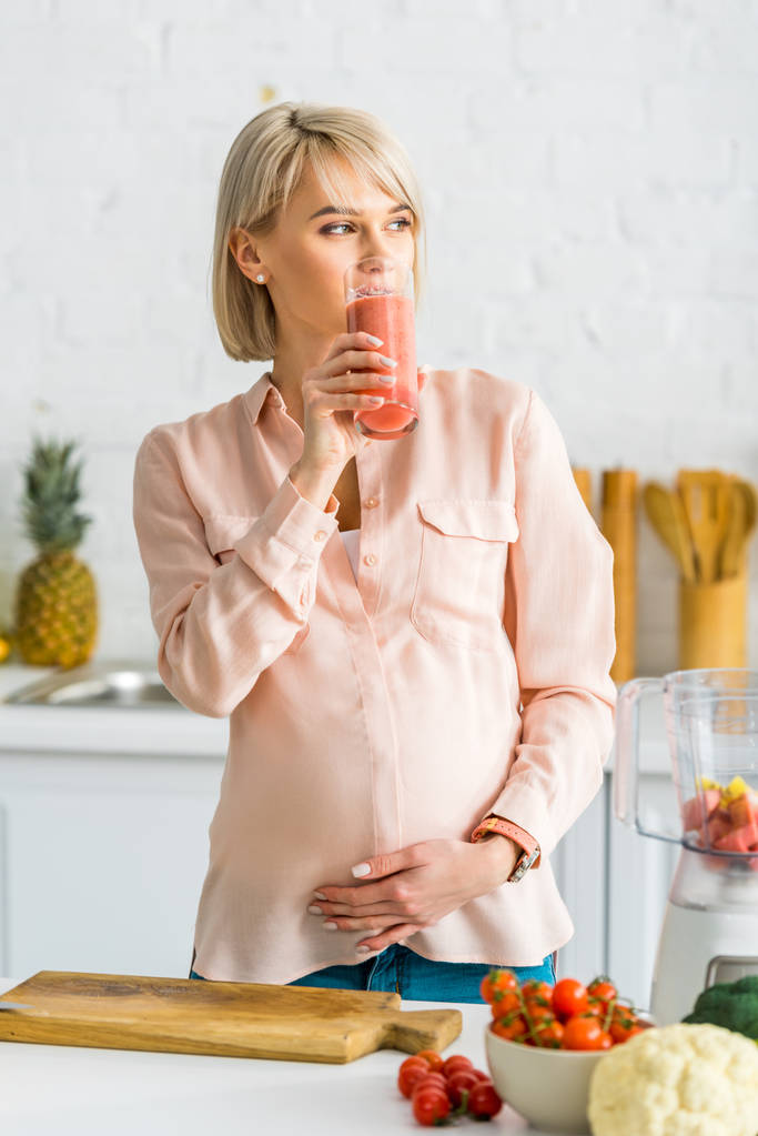 Blonde schwangere Frau trinkt Smoothie in der Nähe von Gemüse in Küche  - Foto, Bild