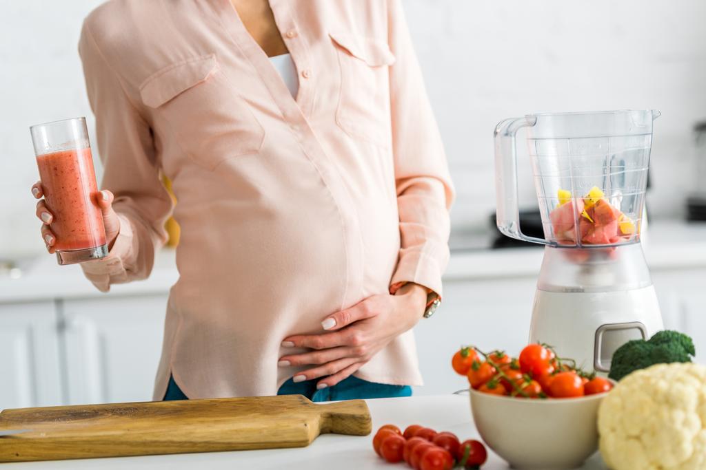 περικομμένη θέα της εγκύου γυναίκα κρατώντας ένα ποτήρι χυμό κοντά στα λαχανικά στην κουζίνα  - Φωτογραφία, εικόνα
