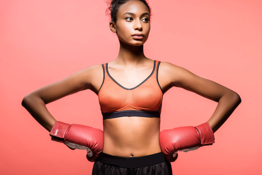 beautiful african american sportswoman in boxing gloves posing with hands akimbo isolated on coral - Photo, Image