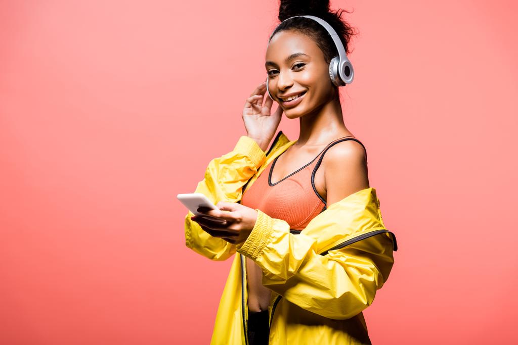 krásné usmívající se africká americká sportovkyně ve sluchátkách při pohledu na fotoaparát a pomocí smartphone izolované na coral - Fotografie, Obrázek