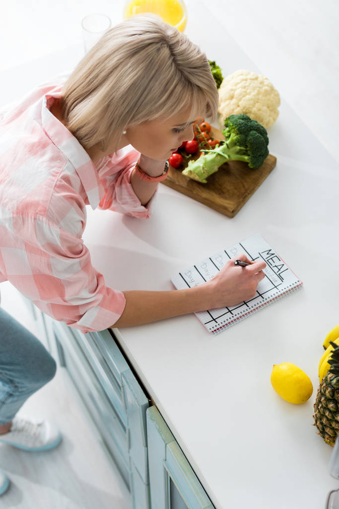 felülről véve a szőke lány írásban notebook-ban étkezési betűkkel közeli összetevők a konyhában - Fotó, kép
