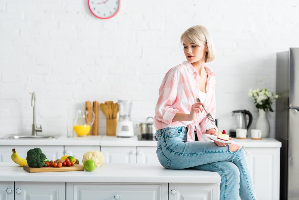 blonde girl holding fork and saucer with sweet cake and looking at organic fruits and vegetables  - Photo, Image